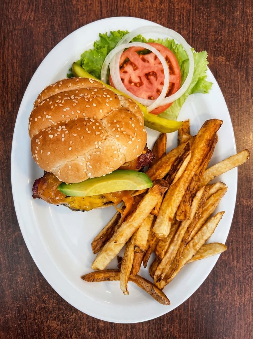 A cheeseburger with avocado, lettuce, tomato, onion, and pickles, served with a side of fries.