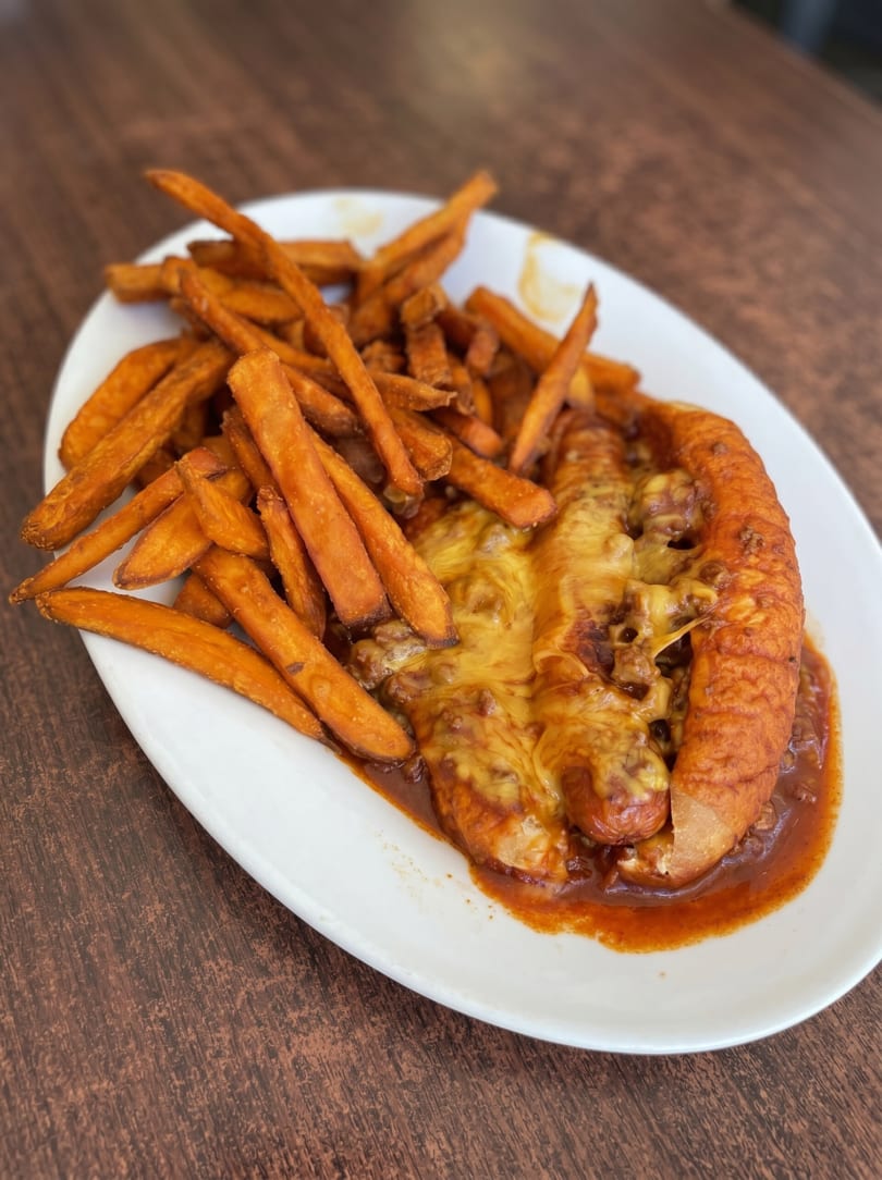 Chili cheese dog with sweet potato fries on a white plate, on a wooden table.